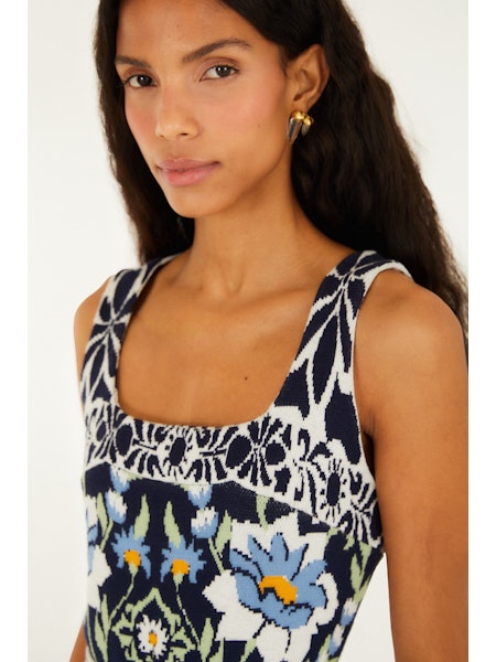 A woman with long dark hair models the Zertz MANOELA KNIT MIDI DRESS, a square-neck knit dress with a blue, white, and yellow floral print. She pairs it with gold hoop earrings and poses neutrally against a plain backdrop.