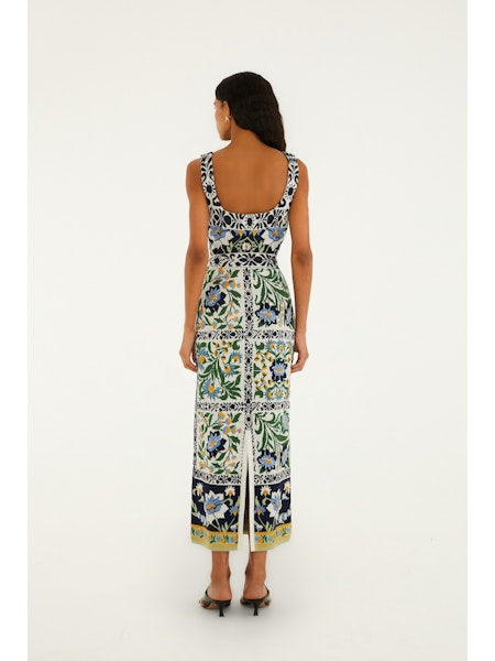 A woman with long dark hair faces away, wearing the Zertz MANOELA KNIT MIDI DRESS—a sleeveless, breathable dress with colorful floral and geometric patterns. She pairs it with heels against a plain white background.