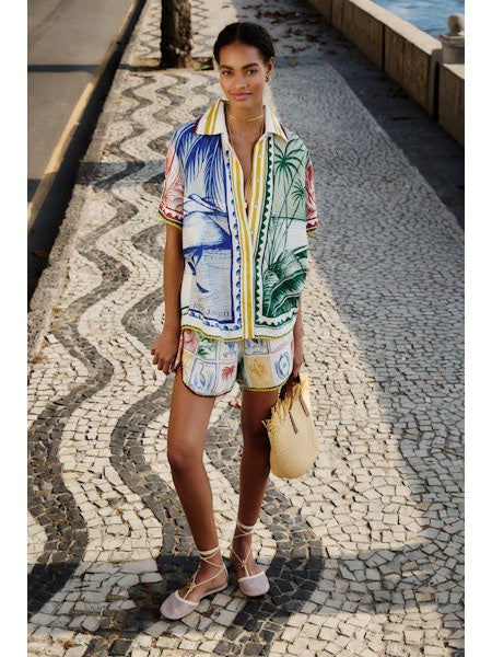 A woman stands on a patterned stone sidewalk by the water, wearing Zertz's BRASIL VINTAGE SHORTS with a relaxed fit, a colorful tropical-print shirt, lace-up sandals, and holding a straw bag. A road and stone wall appear in the background.