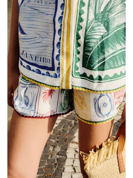 Wearing Zertz BRASIL VINTAGE SHORTS and matching tropical postage stamp top, a person stands on cobblestones. The relaxed outfit is paired with a fringed woven straw bag; only their lower half is visible.