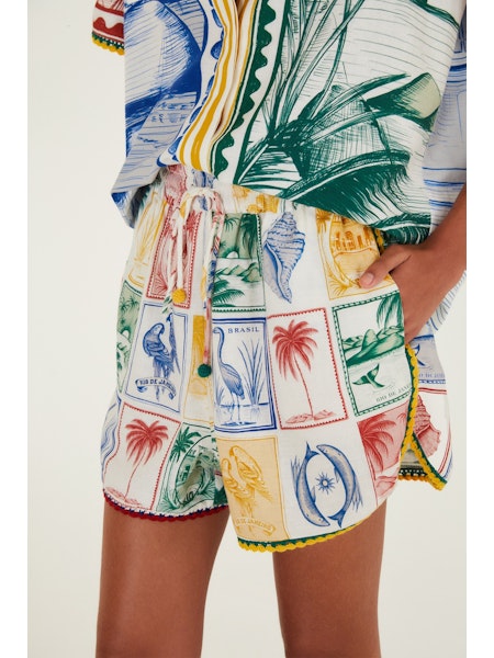 A person wears Zertz BRASIL VINTAGE SHORTS with a matching tropical print shirt featuring palm trees, birds, seashells, and Brazilian motifs in bold red, yellow, green, and blue. The retro look is finished with a hand casually in their pocket.
