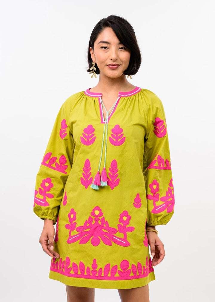 A woman with short dark hair smiles in the Zertz APPLIQUE TUNIC—a relaxed, long-sleeve yellow-green top with bright pink floral patterns, a keyhole neckline, and blue tassels—standing against a plain white background.