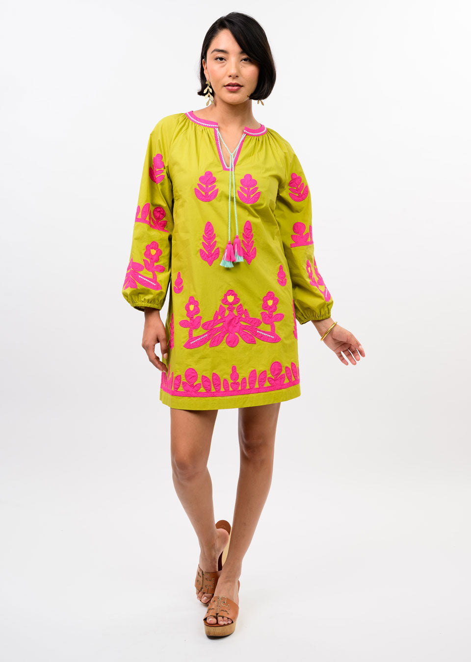A woman poses against a white background wearing the Zertz APPLIQUE TUNIC—a lime green tunic with bright pink floral embroidery, long sleeves, and neckline tassels—showcasing its relaxed silhouette.