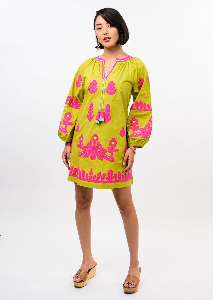 A woman models the Zertz APPLIQUE TUNIC in lime green with bright pink floral embroidery, a tassel-tie neckline, and tan platform sandals, highlighting its relaxed silhouette against a plain white background.