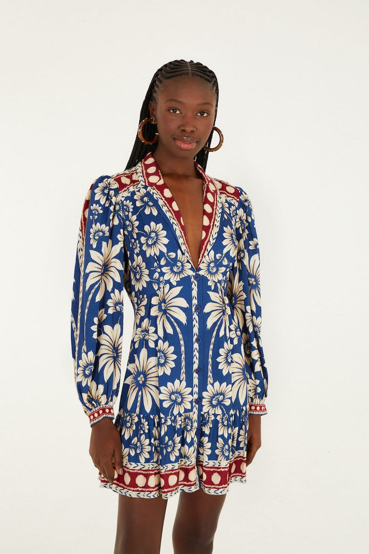 A woman wears the Zertz LUNA BLUE LG SLV MINI DRESS, featuring white floral patterns and red accents, standing against a plain white background and smiling with her hand by her side.