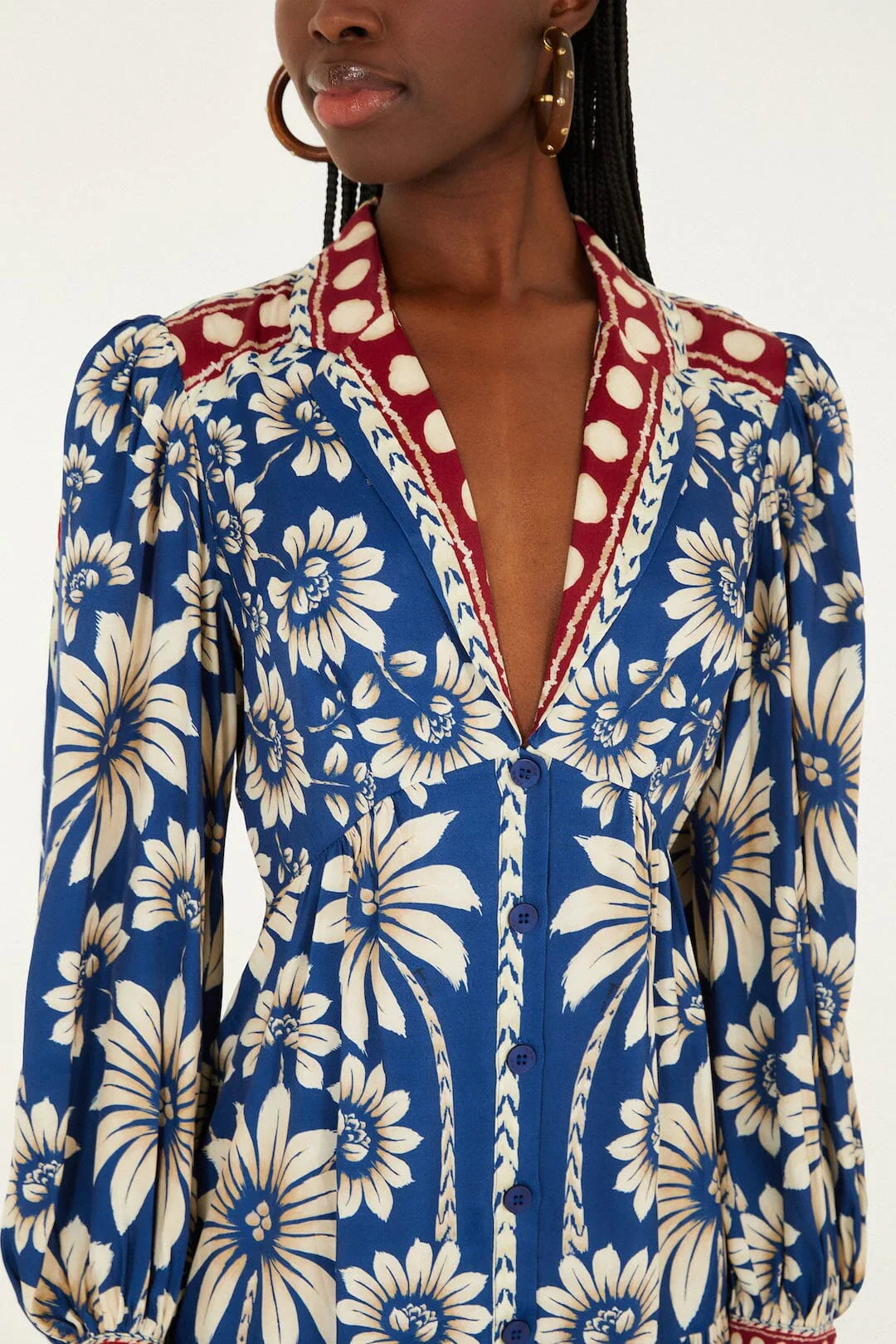 A person wears the Zertz LUNA BLUE LG SLV MINI DRESS with large cream floral patterns, puffed long sleeves, and a deep V-neck with a maroon and white polka dot collar, styled with hoop earrings and braided hair.