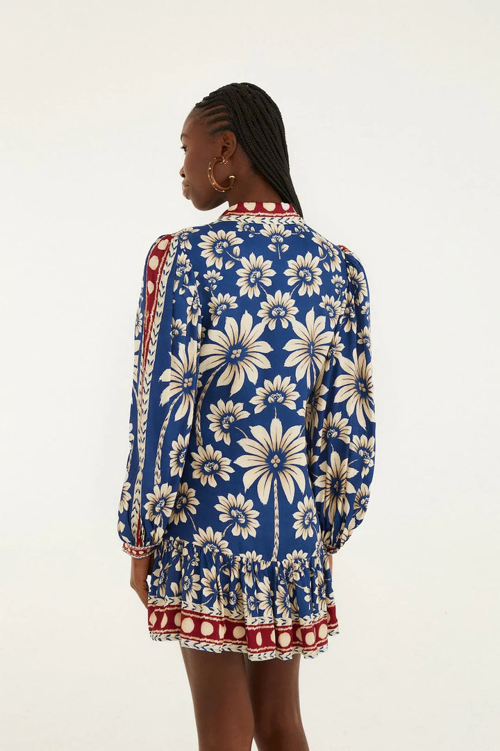 A woman with braided hair wears the Zertz LUNA BLUE LG SLV MINI DRESS, featuring long sleeves and bold white and yellow florals with red accents. She stands facing away from the camera against a plain background.
