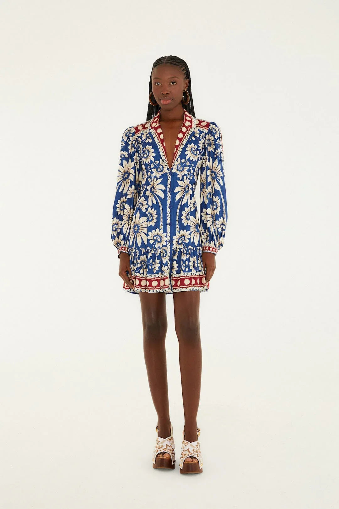 A woman stands against a plain background wearing the LUNA BLUE LG SLV MINI DRESS by Zertz, featuring bold white floral patterns, red accents, long puff sleeves, and white platform sandals.