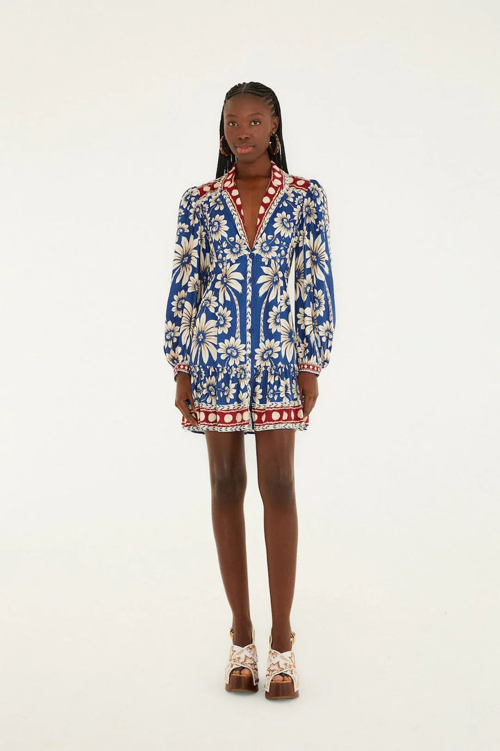 A woman stands against a plain background wearing the LUNA BLUE LG SLV MINI DRESS by Zertz, featuring bold white floral patterns, red accents, long puff sleeves, and white platform sandals.