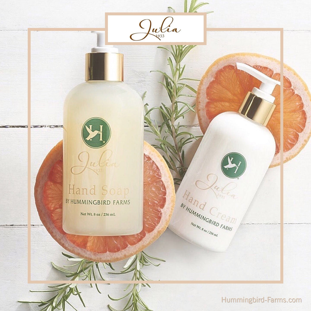 The Julia 1933 Hand Soap &amp; Cream Duo by Hummingbird Farms is pictured with rosemary sprigs and grapefruit slices. The bottles feature a green hummingbird logo on a light background.