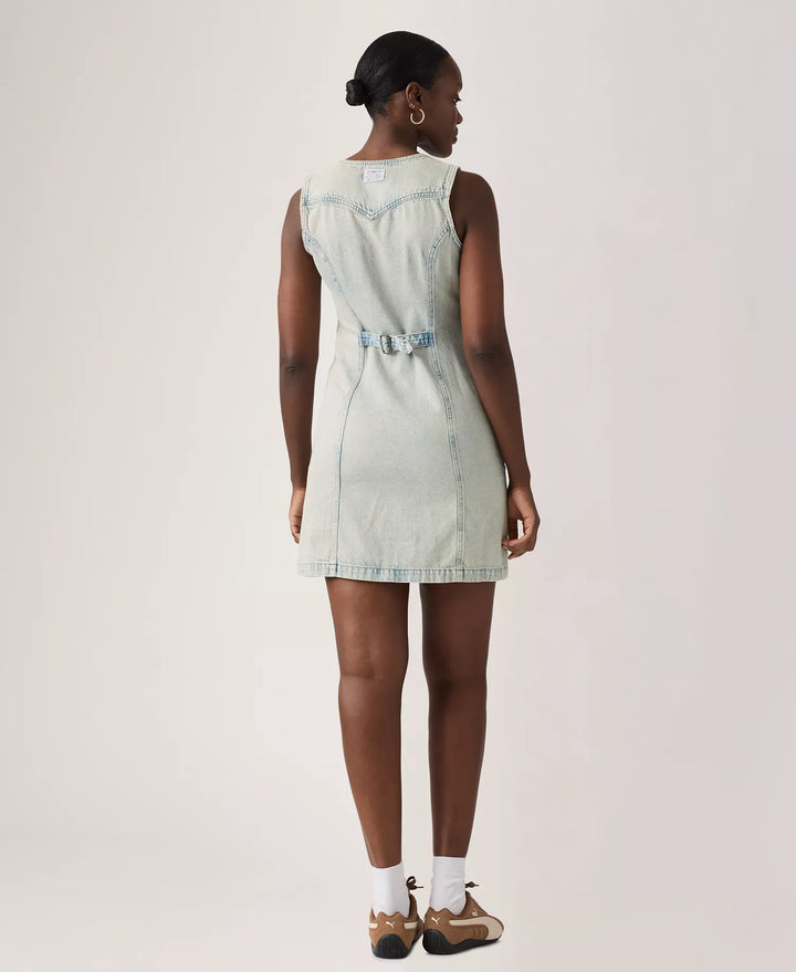 A woman faces away, wearing the Zertz VIANNA VEST DRESS- A LITTLE HAZY DRESS in light denim with a back belt detail, paired with white socks and brown shoes, set against a plain light background.
