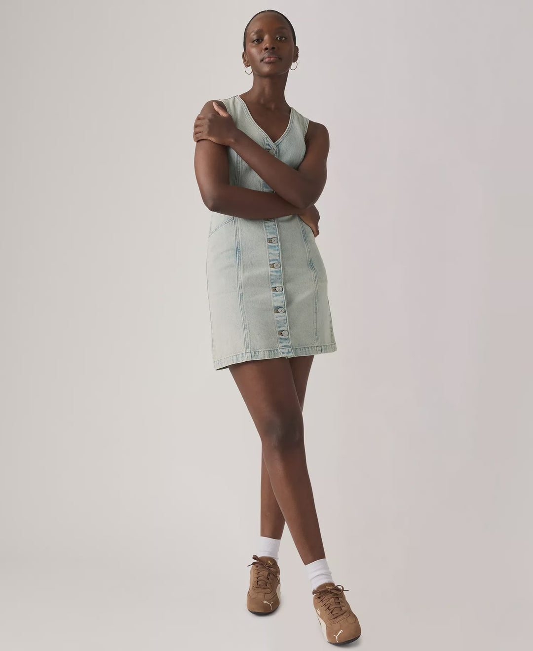 A person stands with arms crossed, wearing the Zertz VIANNA VEST DRESS- A LITTLE HAZY DRESS, paired with white socks and brown sneakers, against a plain light background.