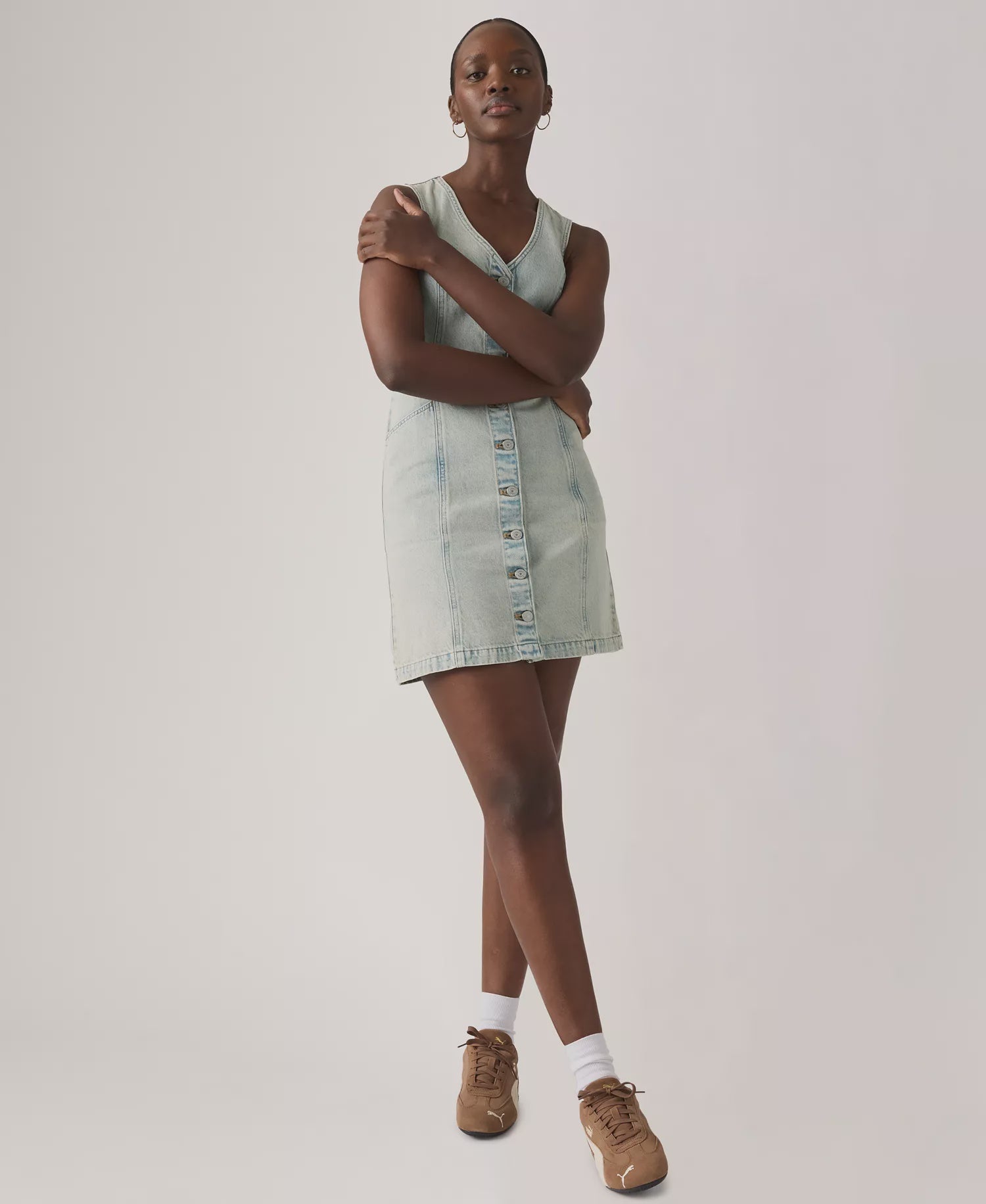 A person stands with arms crossed, wearing the Zertz VIANNA VEST DRESS- A LITTLE HAZY DRESS, paired with white socks and brown sneakers, against a plain light background.