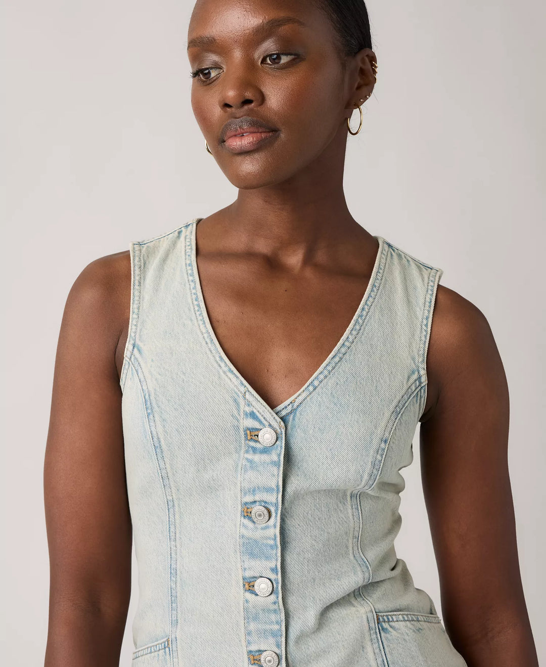 A woman wears the Zertz VIANNA VEST DRESS- A LITTLE HAZY DRESS, a light wash, sleeveless denim dress with a V-neck and button-down front. She stands against a plain light background, styled with short hair and gold hoop earrings.