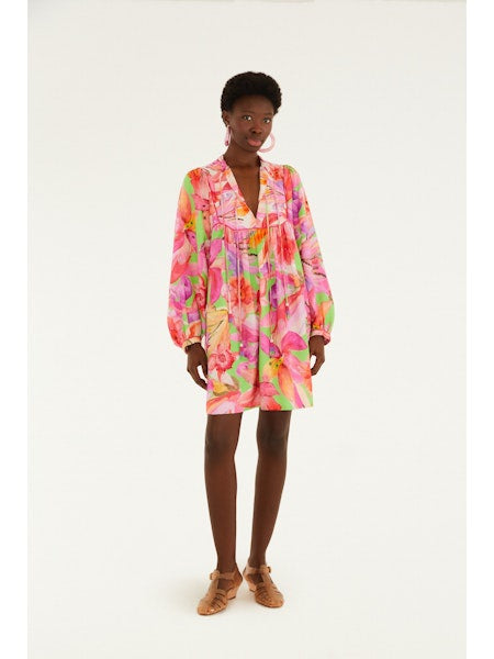 A woman poses against a white background wearing the Zertz ARTSY FLORA GREEN MINI DRESS, a loose, long-sleeved mini dress featuring pink, orange, and green botanical prints, styled with brown flat sandals and hoop earrings.