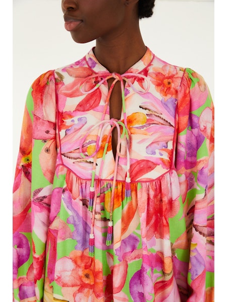 A person wears the Zertz ARTSY FLORA GREEN MINI DRESS featuring pink, red, purple, and green botanical prints. The keyhole neckline ties in bows and billowy sleeves add flair. The person’s face is partially visible.
