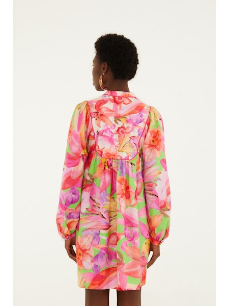 A person with short curly hair is seen from behind wearing the Zertz ARTSY FLORA GREEN MINI DRESS, a long-sleeve piece featuring vibrant pink, orange, and green botanical prints against a plain white background.