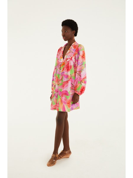 A woman poses in a studio against a white background, wearing the Zertz ARTSY FLORA GREEN MINI DRESS—a colorful, pink and green floral mini with long sleeves and a v-neckline—and tan sandals, her legs stylishly crossed.