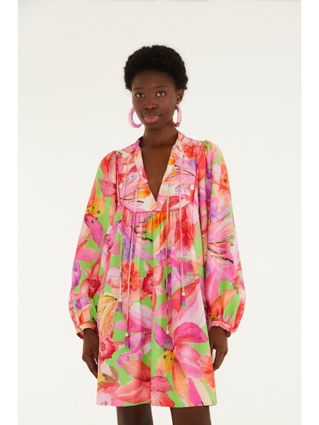 A woman models the Zertz ARTSY FLORA GREEN MINI DRESS, showcasing vibrant pink, orange, and green botanical prints against a white background. She completes the look with large pink hoop earrings.