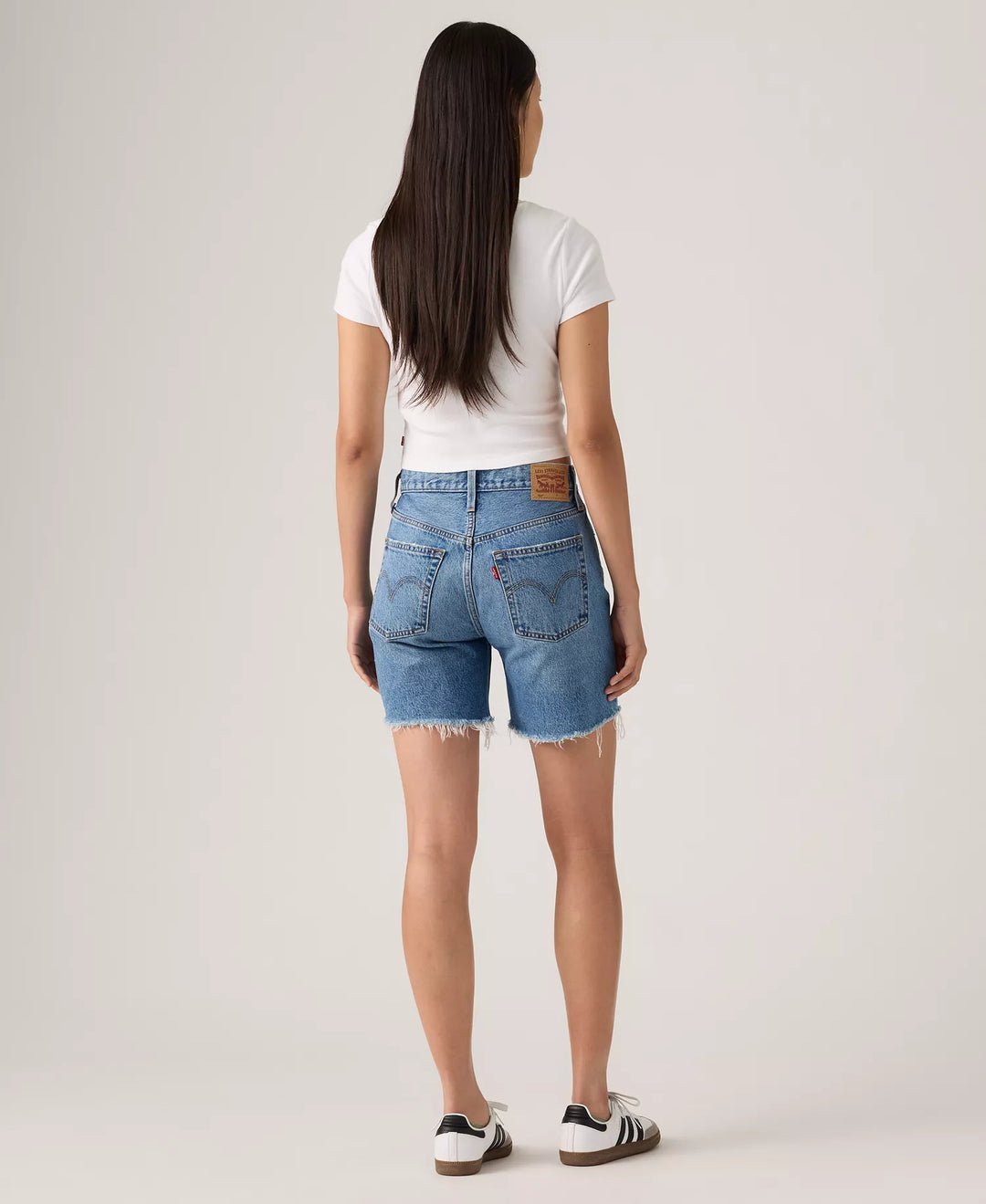 A woman faces away, wearing a vintage-style white tee, Zertz 501 MID THIGH SHORTS - BLOW BIG HORNS in blue with frayed hems, and white sneakers with black stripes, shown against a plain light background.