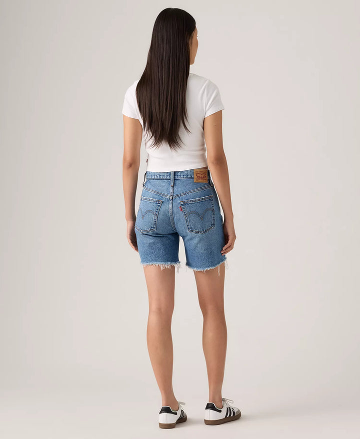 A woman faces away, wearing a vintage-style white tee, Zertz 501 MID THIGH SHORTS - BLOW BIG HORNS in blue with frayed hems, and white sneakers with black stripes, shown against a plain light background.