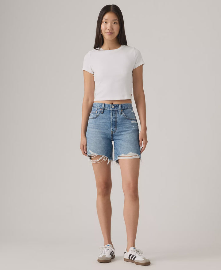 A woman faces forward against a light gray background, wearing a white cropped t-shirt, Zertz 501 MID THIGH SHORTS - BLOW BIG HORNS, and white sneakers with black stripes.