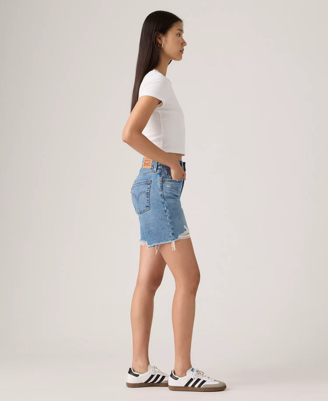A woman stands in profile against a plain background, wearing a white fitted t-shirt, Zertz 501 MID THIGH SHORTS - BLOW BIG HORNS, and white Adidas sneakers with black stripes. Her hands are in her shorts pockets and her hair is straight and loose.