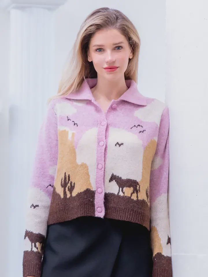 A woman with long blonde hair wears the Zertz JESSIE HORSE KNIT SWEATER in light purple, featuring a desert scene with cacti, horses, and birds. She pairs it with a black skirt and stands against a white background.