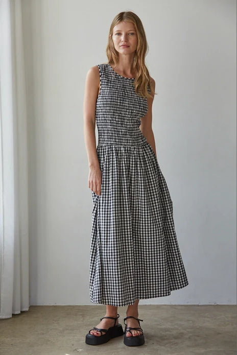 A woman with long, wavy hair wears the Zertz SAYLER GINGHAM SMOCKED DRESS and black platform sandals indoors, smiling slightly with one hand by her side. Soft white curtains appear on the left.