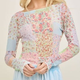 A woman wears the Picnic Patch Mesh Top by Loca on Main, a long-sleeve shirt with colorful floral patchwork, paired with light blue high-waisted pants. Photo shows her from shoulders to waist.