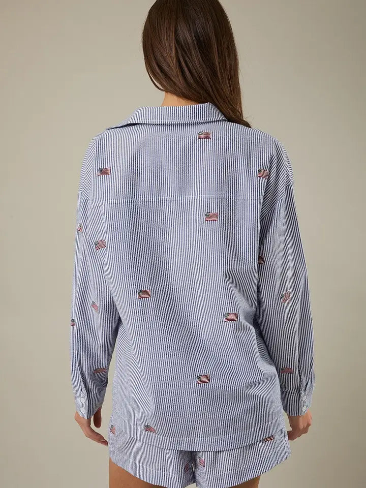 A person with long brown hair is shown from behind wearing the Zertz STRIPE FLAG BUTTON DOWN TOP, featuring blue and white stripes with small red designs. The classic collar shirt has long sleeves and is paired with matching shorts against a plain background.