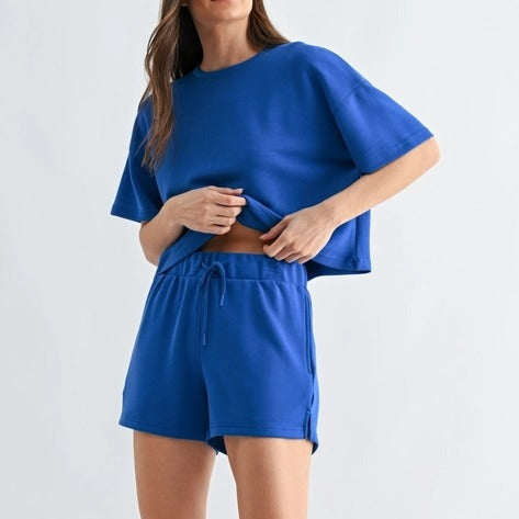 A woman wearing the Loca on Main Laguna Classic Set stands against a plain light background, slightly lifting her shirt to reveal her midriff.