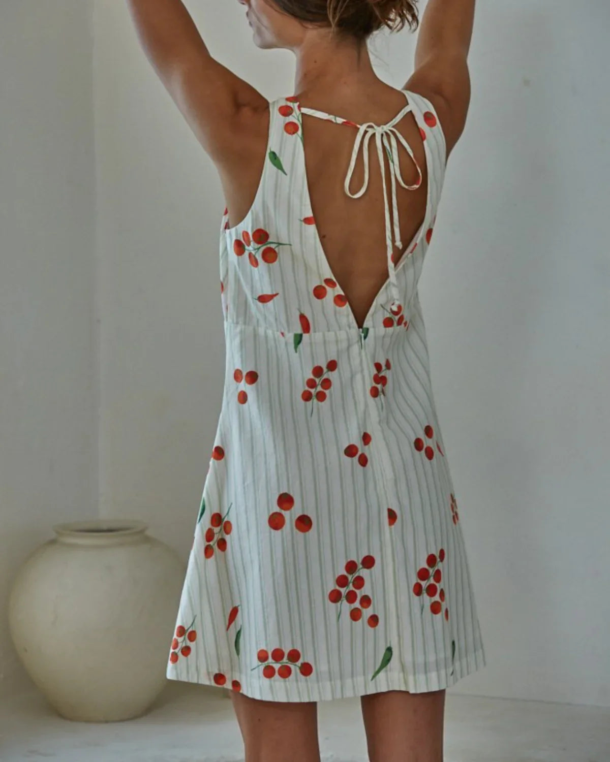 A woman with raised arms models the Zertz CAPRESE MINI DRESS, a sleeveless, backless white mini dress with vertical stripes and cherry patterns tied at the upper back. A light ceramic pot rests on the floor nearby.