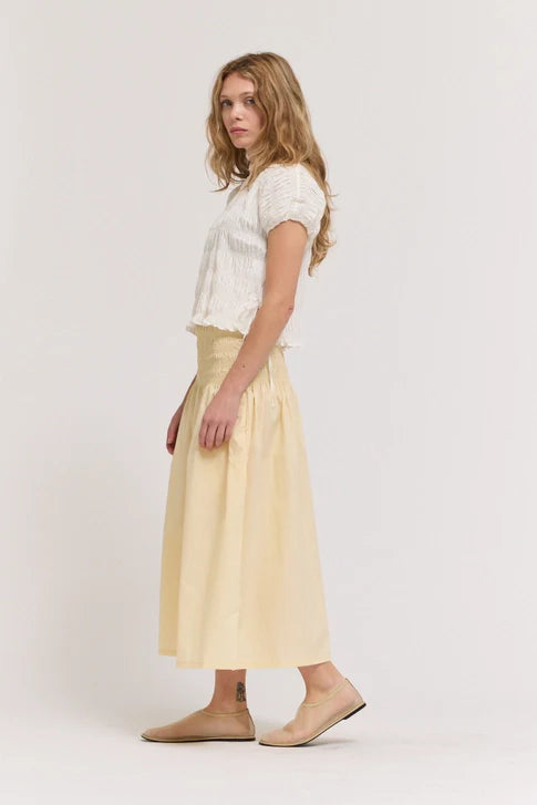 A woman with wavy hair wears the Zertz SMOCKED WAIST SKIRT, a high-waisted light beige piece, paired with a short-sleeve white textured top. She stands sideways against a plain background, highlighting this essential skirt.