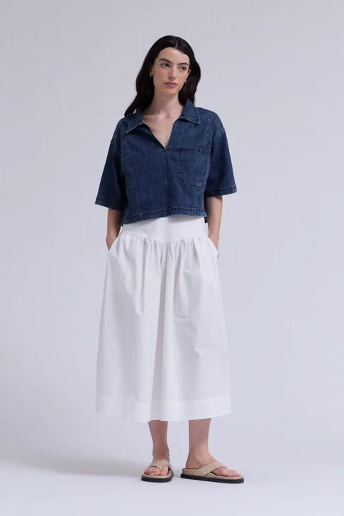 A woman models the Zertz DENIM CROP SHIRT over a white, ankle-length skirt with pockets. With long dark hair and tan sandals, she stands against a plain background, hands tucked casually in her skirt pockets.