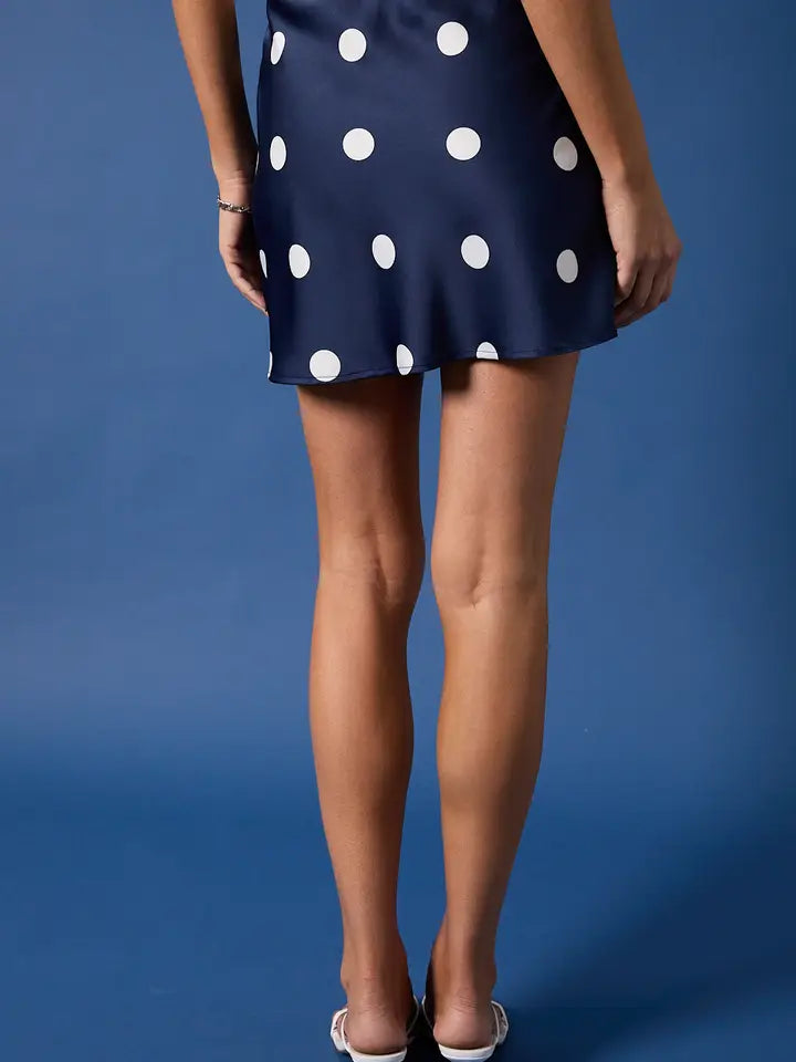 A person faces away against a plain blue background, wearing the Zertz MINI SATIN SKIRT featuring large white polka dots and paired with white heeled shoes.