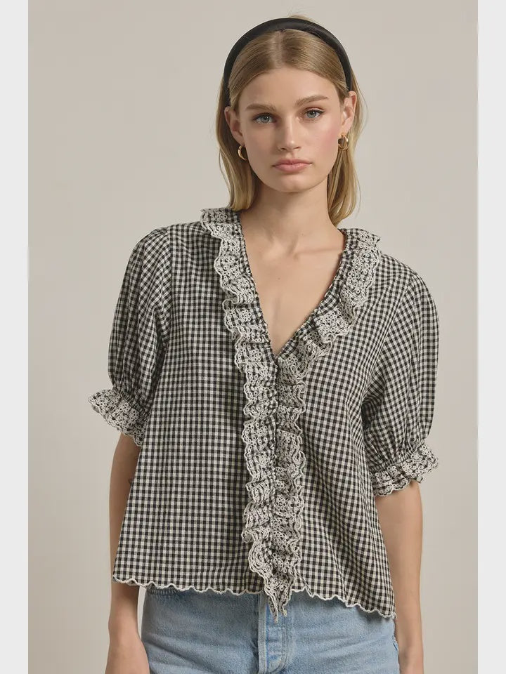 A woman wears the Zertz GINGHAM LACE RUFFLE BLOUSE—vintage-inspired with black and white checks, lace ruffles, puffed sleeves, and a scalloped hem—paired with light blue jeans, straight blonde hair, a black headband, and hoop earrings.