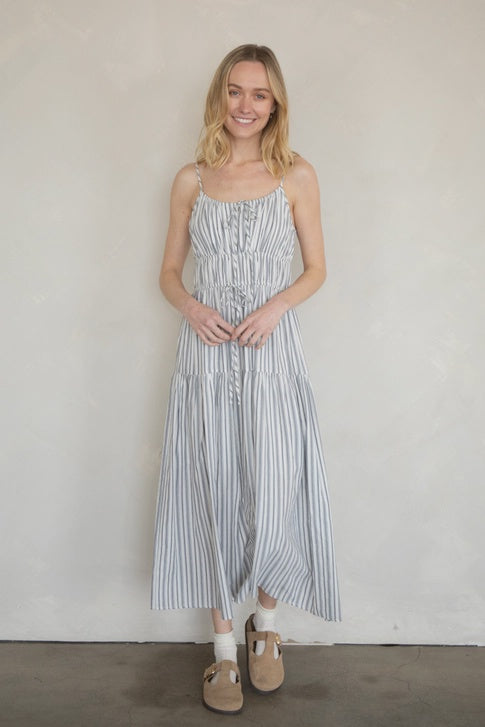 A woman with blonde hair wears the Zertz MAXI COTTON DRESS, a casual sleeveless striped style, with tan sandals and white socks, standing and smiling against a plain light-colored wall.