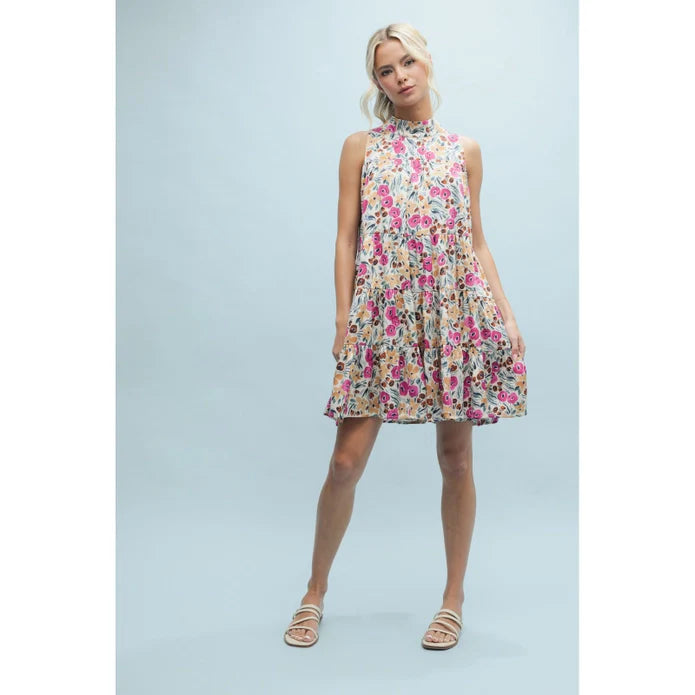 A woman models the Zertz SLVLESS PATTERN DRESS, a sleeveless mini dress with a high neckline, floral print, ruffled hem, and A-line silhouette. She pairs it with flat sandals against a plain light blue background.