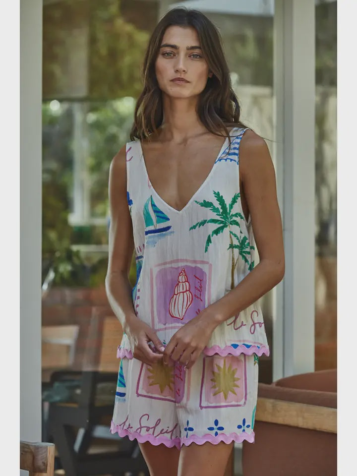 A woman stands indoors near a window in Zertz’s MONTMARTRE WOVEN SHORTS, styled with a sleeveless white dress featuring a deep V-neck and colorful tropical prints for an effortlessly breezy, relaxed look.