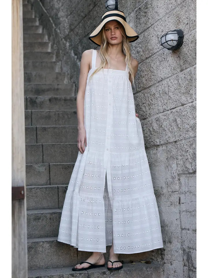 A woman stands on stone stairs wearing the Zertz EYELET BUTTON FRONT MAXI DRESS, a sleeveless white summer dress, paired with a beige sun hat and black flip-flops beside a stone wall with a light fixture.