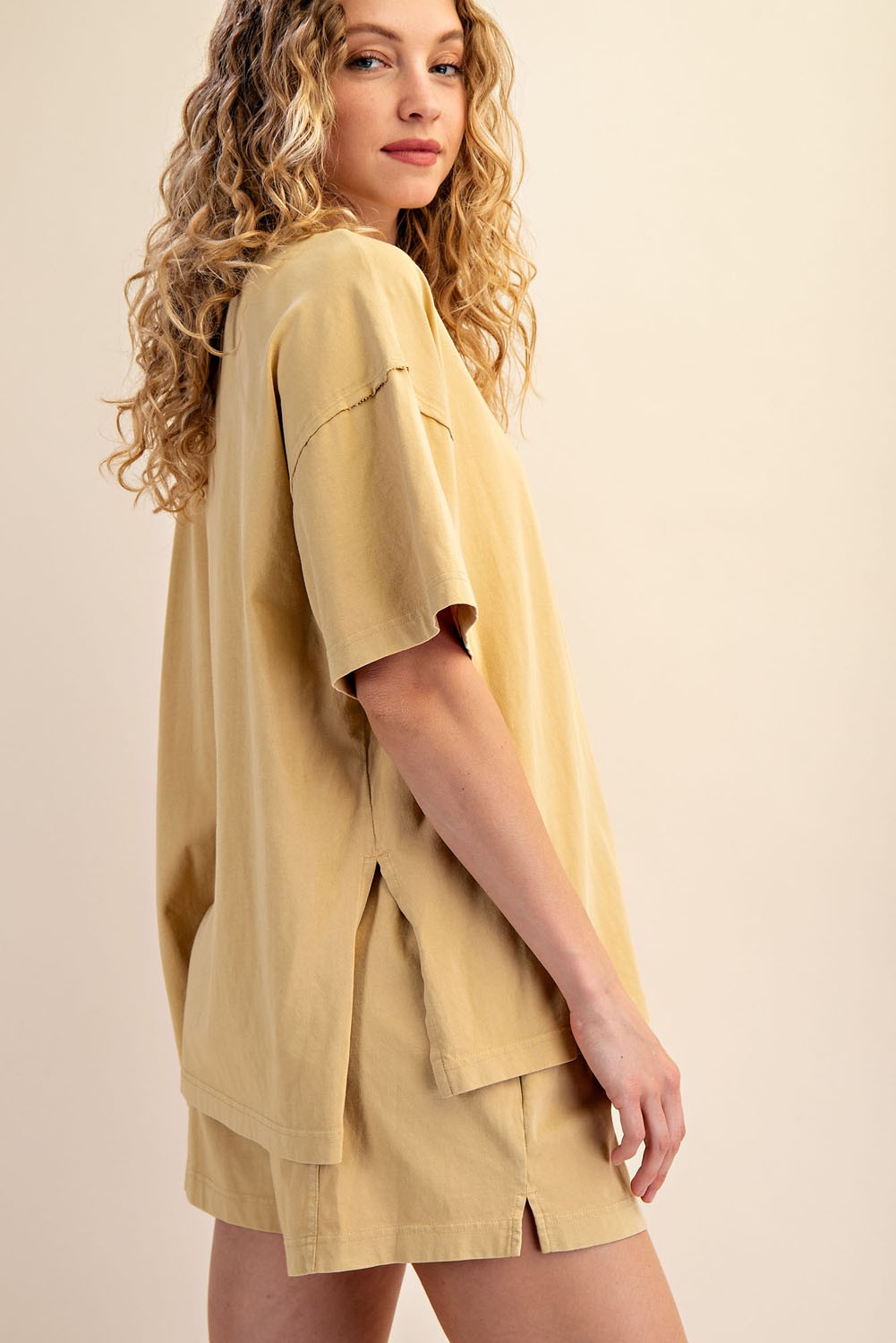 A person with long, curly blonde hair models The Mellow Yellow Set by Loca on Main. This light beige lounge set is loose-fitting and comfortable as they stand sideways, looking toward the camera against a neutral background.