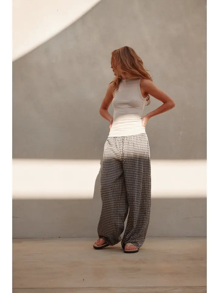 A woman with long wavy hair stands against a plain wall in sunlight, wearing the Zertz SHEER TANK TOP in beige, loose checkered pants, and sandals. She looks to the side with her hands on her hips as light patterns fall on the wall and floor.