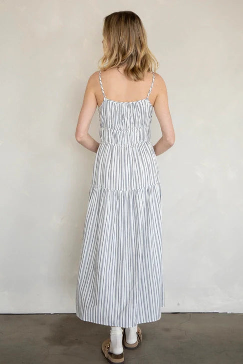 A woman with blonde hair faces away, wearing the Zertz MAXI COTTON DRESS featuring thin blue stripes, paired with tan sandals and white socks, standing on a gray floor against a light beige wall.