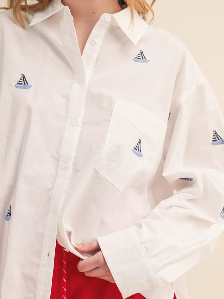 A person wears the Zertz BOAT EMB TOP—an oversized white button-up shirt with blue sailboat patterns and a chest pocket—paired with red pants. The face isn’t visible, making this statement piece the center of attention.