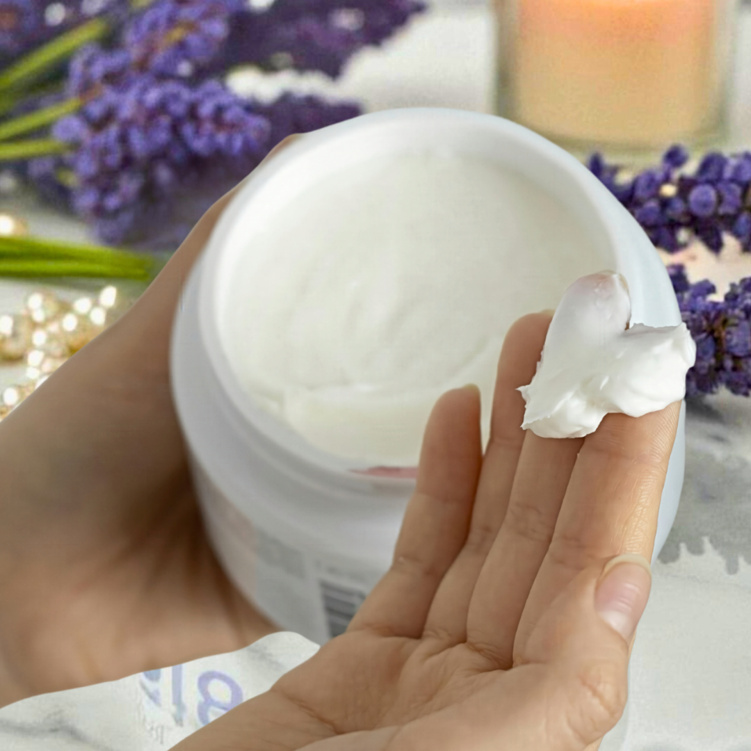 A hand scoops a dollop of Hummingbird Farms Lavender Heavy Cream No.518 from an open jar, as purple lavender flowers, pearls, and a lit candle appear blurred in the background.