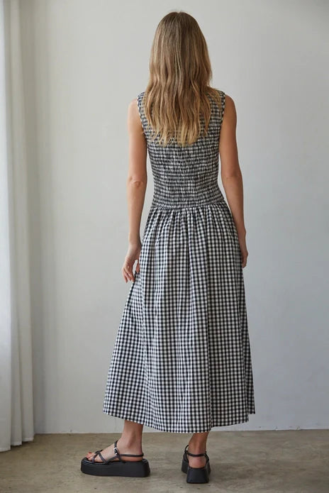 A woman with long, wavy hair stands indoors facing away, wearing the Zertz SAYLER GINGHAM SMOCKED DRESS and black platform sandals near a white wall and window.