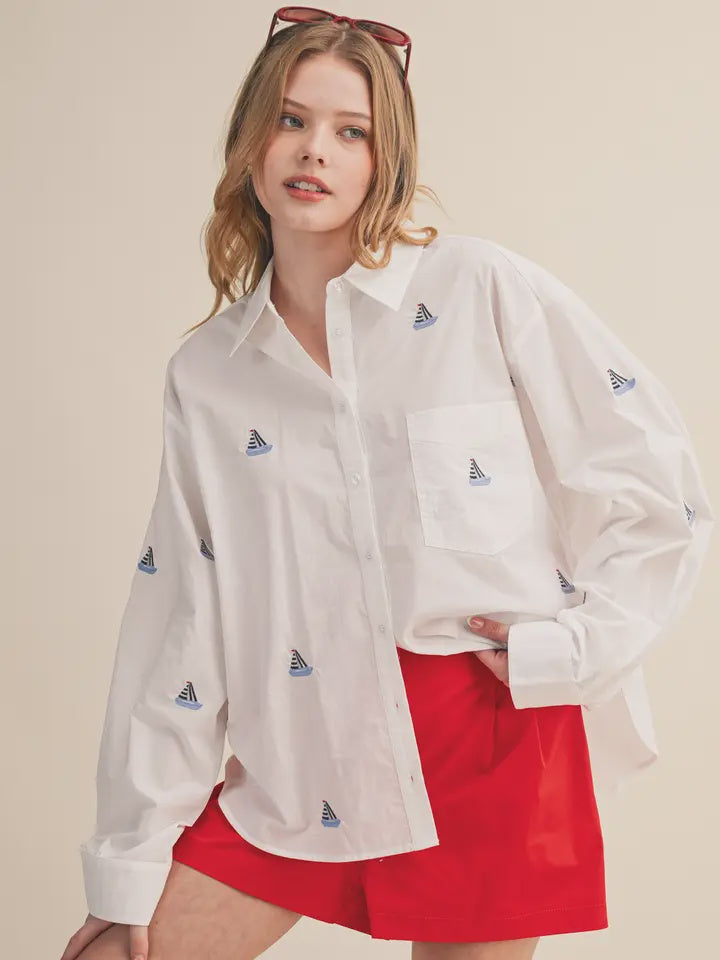 A woman wears the Zertz BOAT EMB TOP, a loose white classic top with small sailboat patterns, paired with red shorts and sunglasses on her head as she poses casually with one hand in her pocket against a neutral background.