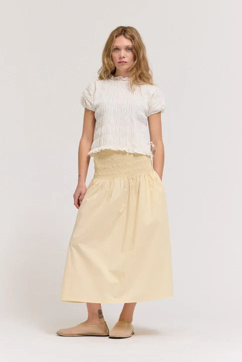 A woman with wavy blonde hair and a neutral expression wears a white textured top and the Zertz SMOCKED WAIST SKIRT in light yellow, paired with beige slip-on shoes—timeless essentials against a plain background.