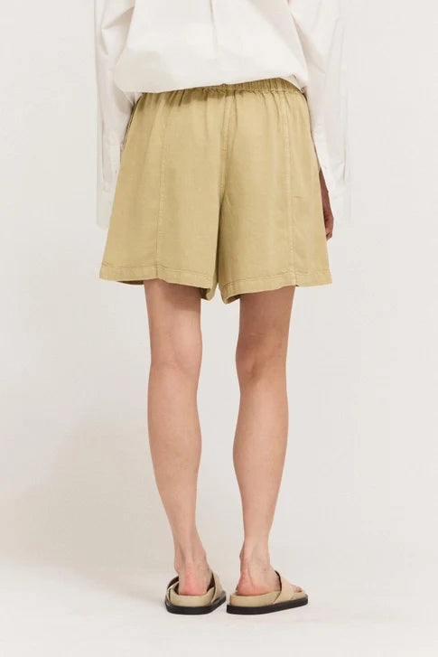 A person stands with their back to the camera, wearing a white long-sleeve shirt, Zertz TENCEL SHORTS in tan with an elastic waistband for comfort, and beige slide sandals against a plain, light background.
